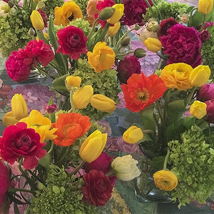 A table top full of little centerpieces of Summer blooms; including yellow tulips, orange poppies and red Ranunculus with green hydrangea for a bridal shower.