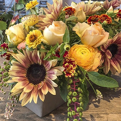This centerpiece arrangement is a wonderful mix of caramel color roses, yellow ranunculus, small zinnias, two tone sunflowers and a bit of pokeberry trailing over the side.
