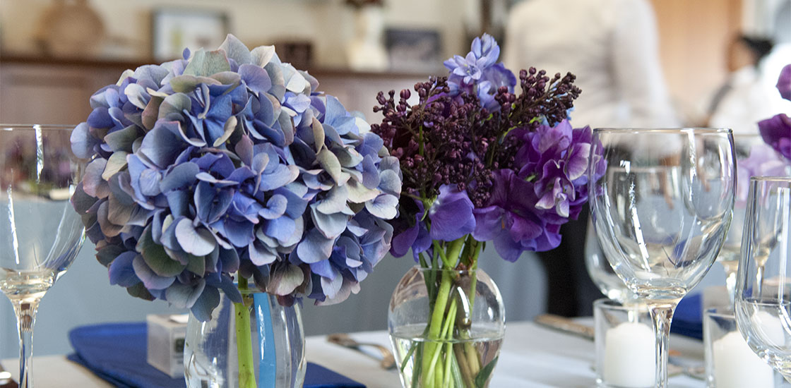A Spring event with the long table decorated with little vases filled with purple and blue Spring flowers, including lilacs with sweet peas to scent the air, mixed with hydrangeas, anemones and hyacinth.